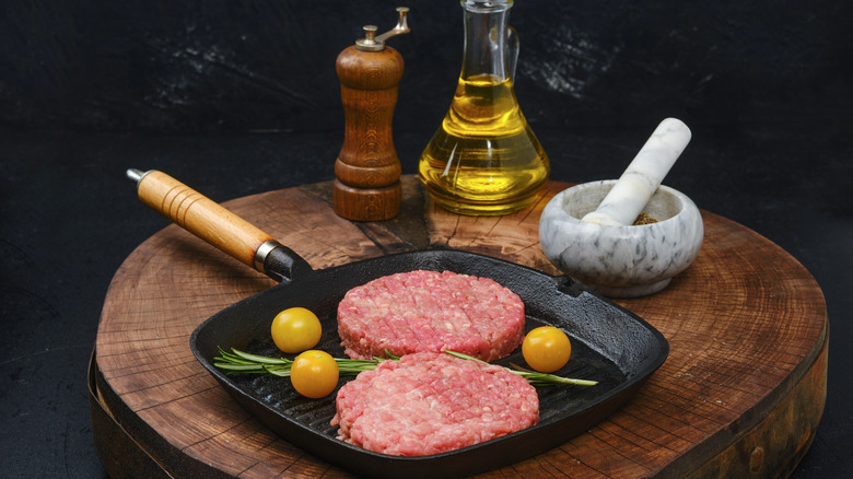 Two burger patties on a cast iron griddle with herbs and cherry tomatoes, on a table next to a peppermill, mortar and pestle, and jug of olive oil