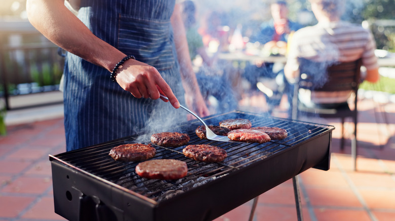 A grillmaster flips burgers on a grill with friends seated at a table in the background