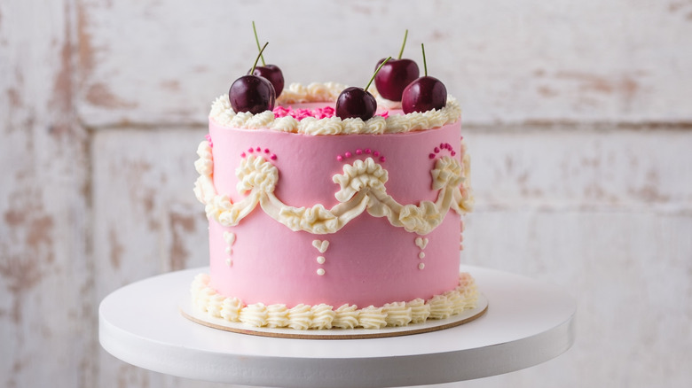 A pink Lambeth cake with cherries on top sits on a white pedestal.