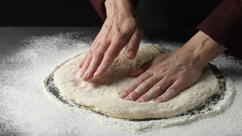 A baker's hands flattens out pizza dough on a table covered in flour