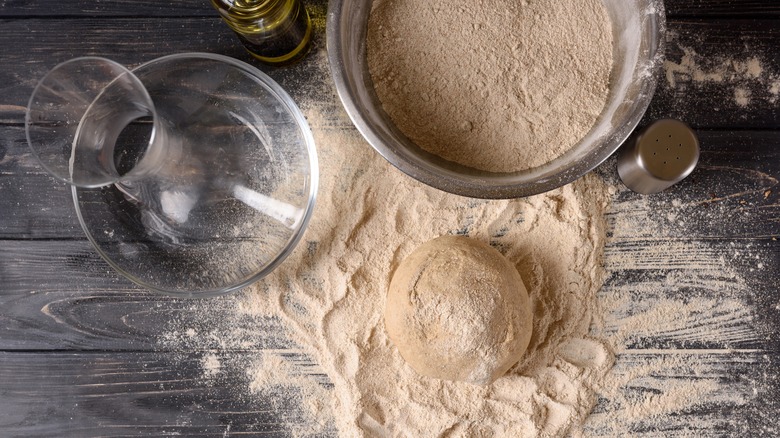 A ball of pizza dough sits on a table next to a bowl of flour and a pitcher of water