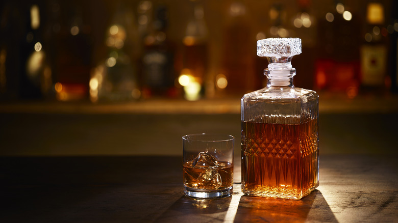 A decanter full of bourbon sits next to a glass with bourbon on the rocks before a well stocked bar