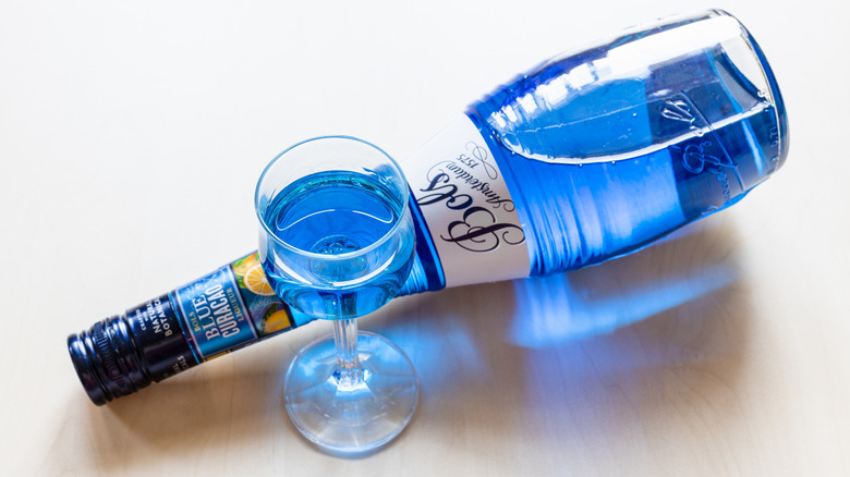 Bottle of blue curacao liqueur laying on its side on a white surface with a wine-style glass half full of the blue liquid in front of it