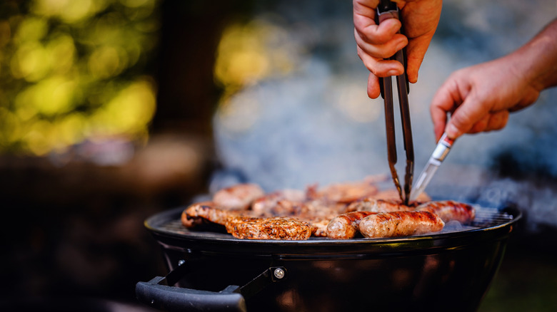 Hands use grill tongs and a meat thermometer on meat cooking over a grill