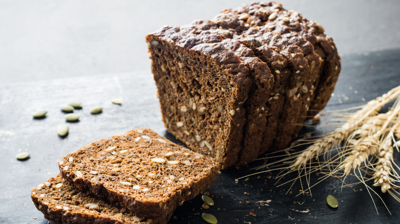 A sliced whole grain rye bread beside grain on a black wooden table