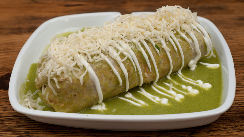 Wet burrito in white plate with green sauce, crema, grated cheese