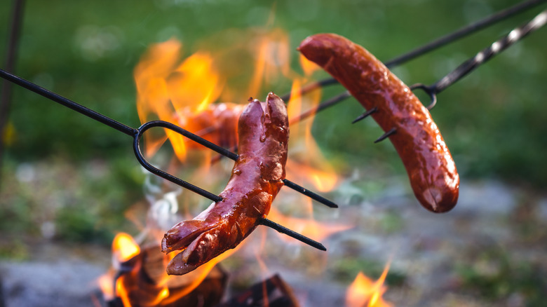 Two sausages being roasted over a campfire with skewers