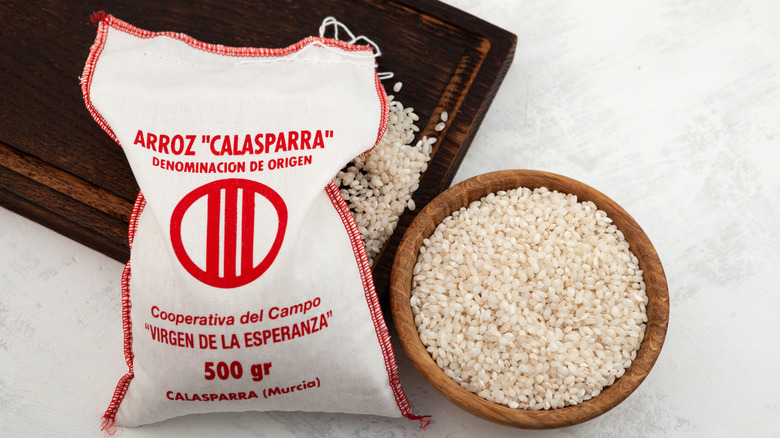 A bag of Calasparra rice spilling grains on a wooden cutting board next to a bowl of uncooked rice