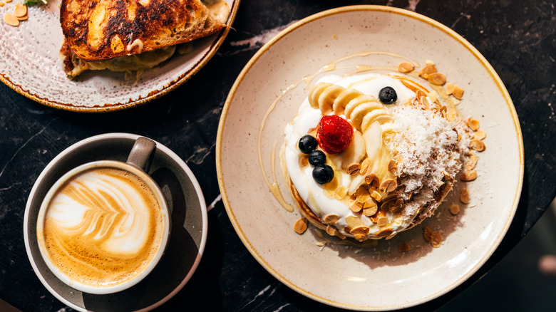 Pancakes topped with whipped cream, oats, bananas and berries sit beside a cup of coffee.