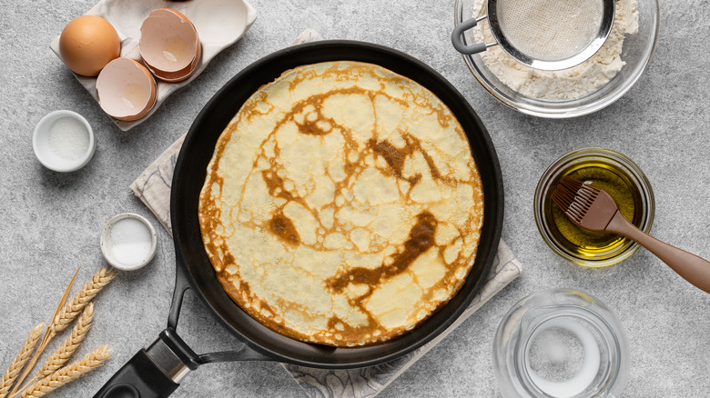 A downward view shows a crepe in a pan alongside neatly cracked eggs, a sifter sitting in flour and a cooking brush in a small cup of oil.