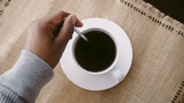 A top-down image of a hand stirring a spoon around a mug full of black coffee