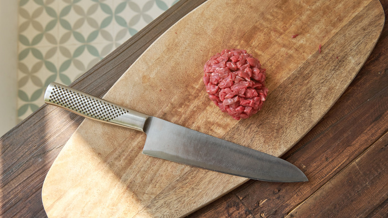 Raw beef cut up for tartare rests next to a sharp knife on a wooden cutting board