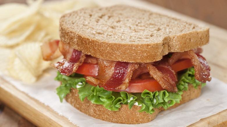 A tasty BLT sandwich sits on a wooden cutting board with potato chips nearby