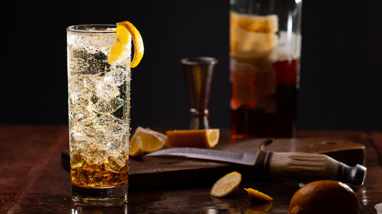 Glass of highball cocktail with ice surrounded by ingredients and a bottle of whiskey