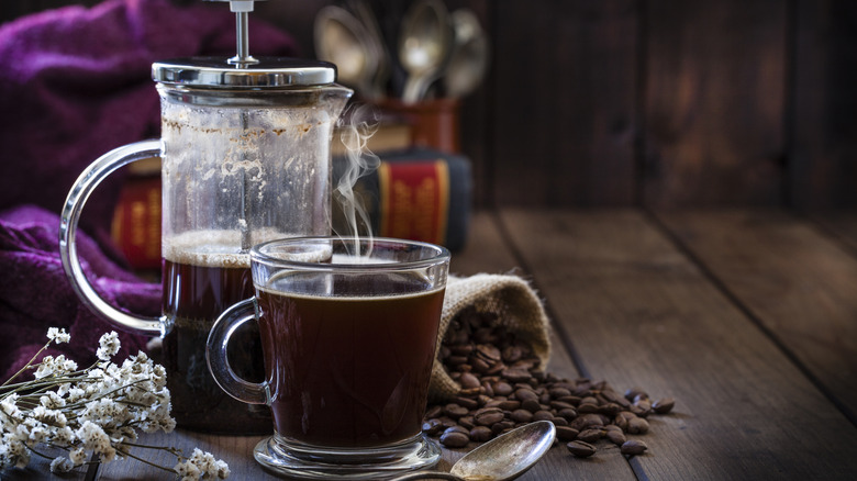 A French press sits, half full, next too a steaming cup of coffee, some baby's breath flowers, and a rustic sack of coffee beans