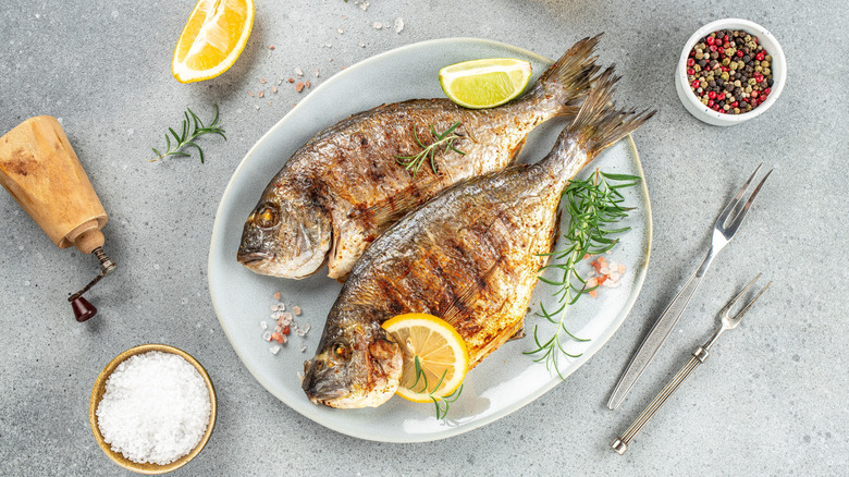 Two whole roasted sea bream on a plate with salt, pepper, lemon, and herbs