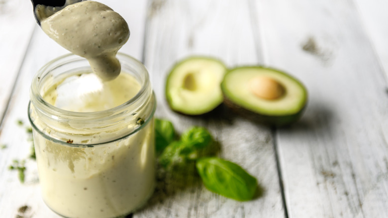 Homemade avocado mayonnaise scooped out of a jar, which sits next to fresh basil and a sliced avocado