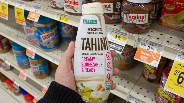 A hand holding a bottle of tahini in front of grocery store nut butter shelf