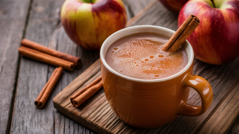 A mug of apple cider is surrounded by apples and cinnamon sticks