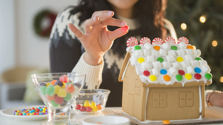 A woman places a red gumdrop on a decorated gingerbread house.