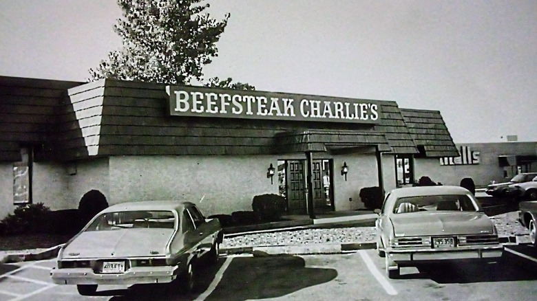 Old fashioned cars sit in a parking lot of a Beefsteak Charlie's location in a black and white photo