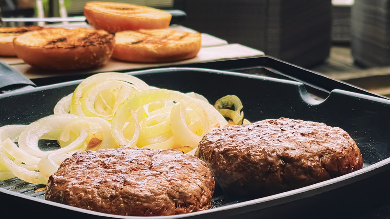 Hamburgers and onions are grilled outdoors in a cast iron pan, with toasted buns sitting nearby