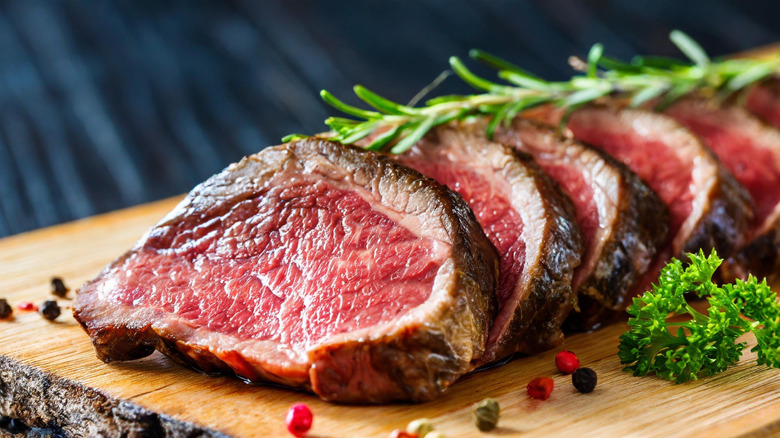 Sliced pieces of rare beef on a wooden surface with a rosemary spring on top and a piece of parsley and colorful peppercorns scattered