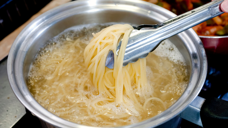 Hand removing pasta noodles from pot of boiling water with metal tongs