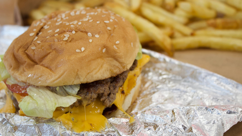 A Five Guys burger sits in the foreground on foil, with fries in the background