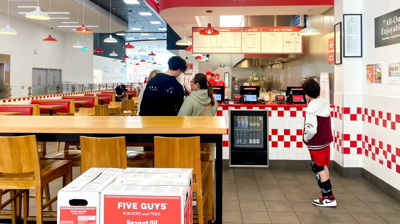 A few people wait in line at a brightly lit Five Guys counter