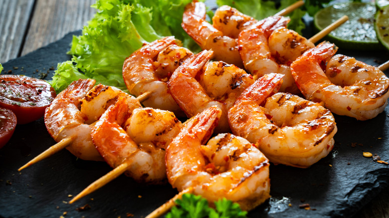 Grilled shrimp on skewers sit on a black slab plate with lettuce and marinated tomatoes