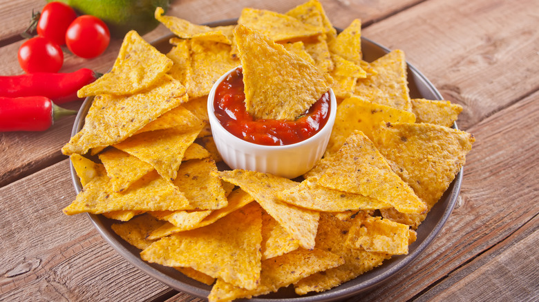 Nacho tortilla chips on a round plate with ramekin of tomato salsa on wood table