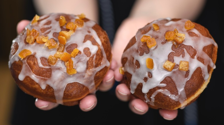 Hands hold out two paczki Polish donuts with sugar glaze and orange peel topping