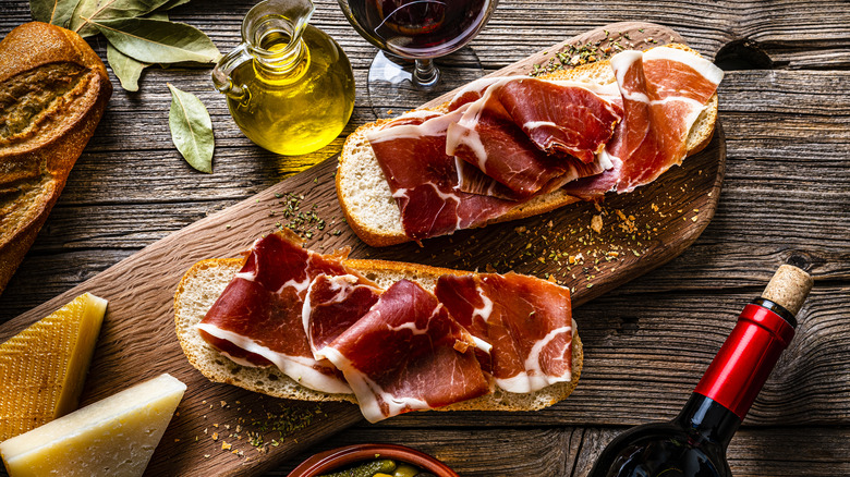 A cutting board with sliced bread covered with jamón Iberíco, surrounded by Manchego cheese, olive oil, a bottle of wine, and a glass of wine.