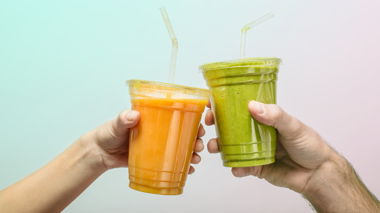 Hands holding plastic cups of green juice and orange-colored juice clink them together.