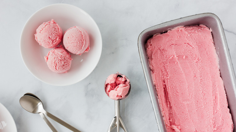 A tub of strawberry ice cream next to bowl and scooper