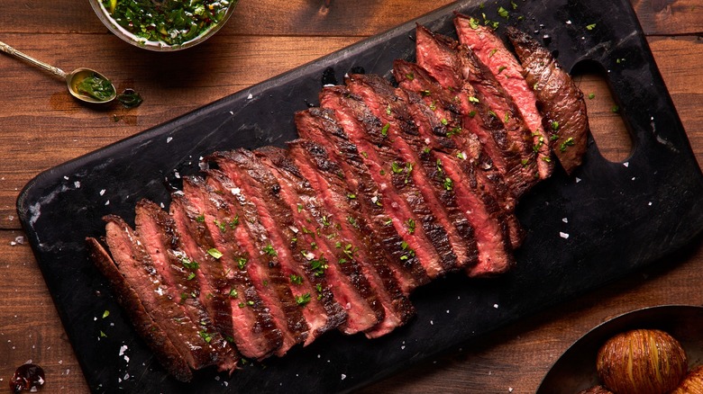 A medium-rare flank steak is served in family-style cuts on a black serving board