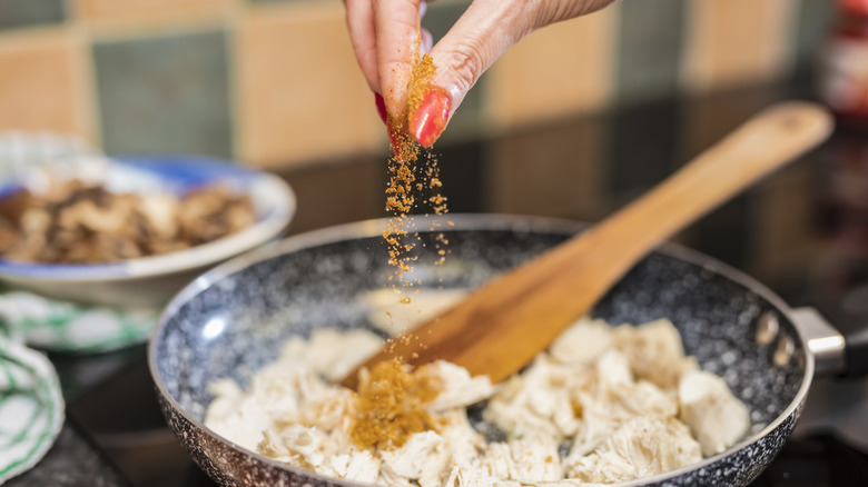 A hand with red nail polish adds seasoning to a chicken dish that sits in a blue speckled bowl