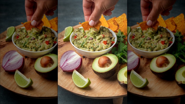 Bowl of guacamole being dipped with a chip displayed with halves of red onion and avocado