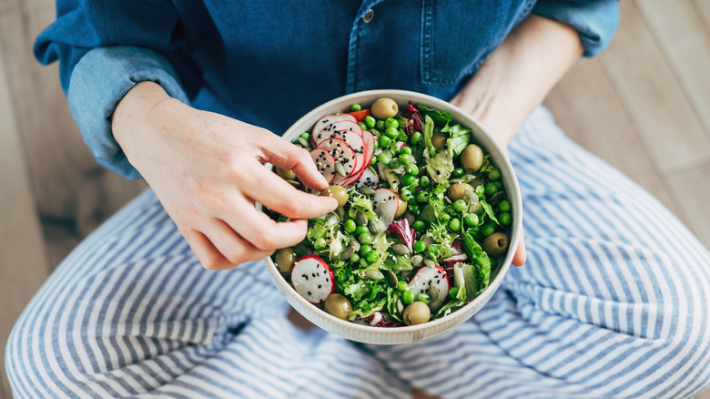 A woman sits cross-legged holding a salad that she is eating with her fingers