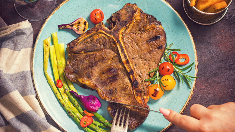 A diner cuts into a well-done T bone steak with a fork and knife