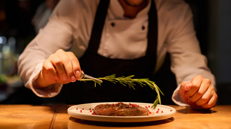 A chef in a white coat and blue apron places a rosemary sprig on a steak