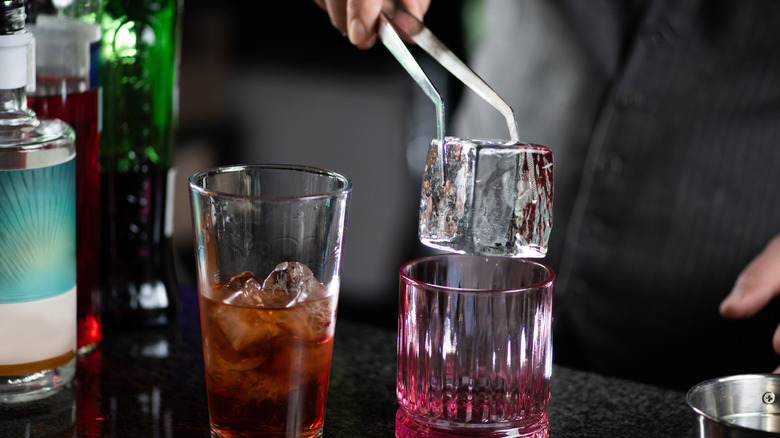 A bartender places a big cube of clear ice in a rocks glass.