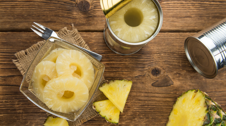 Two cans of pineapple, one open, sit next to a sliced fresh pineapple and a plate full of canned pineapple rings.