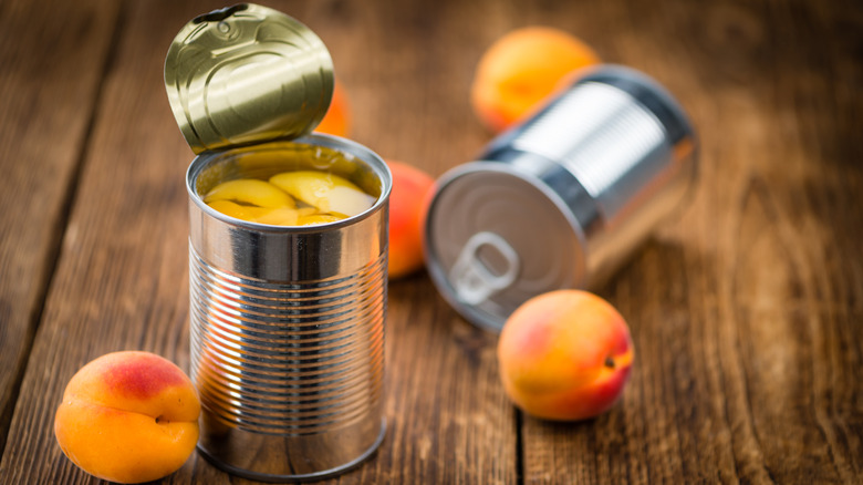 Two cans of peaches, one open, sit on wooden planks next to ripe peaches