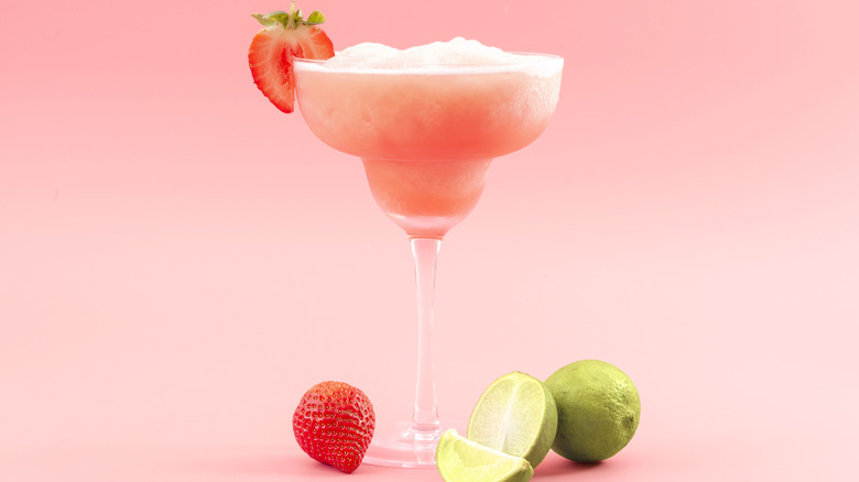 A blended strawberry daiquiri has a fresh strawberry garnish and sits next to a strawberry and sliced limes