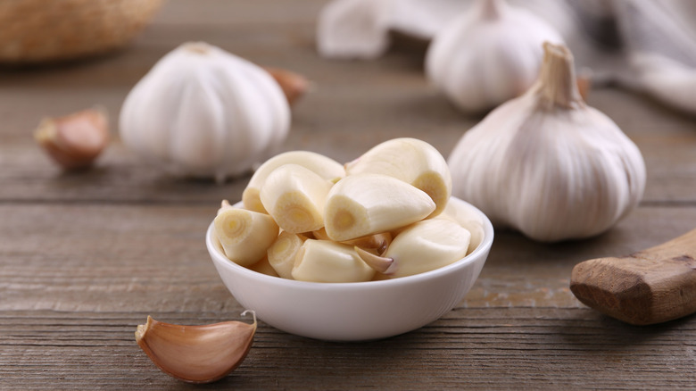 Peeled garlic cloves in a white bowl surrounded by garlic heads