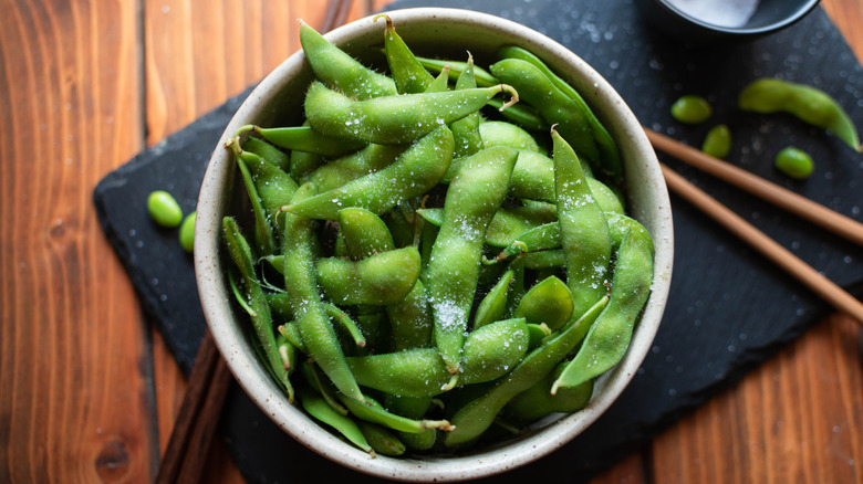 Close up of a bowl of salted edamame sits next to some chopsticks and a bow of salt