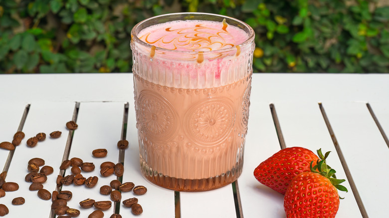 Coffee latte with pink foam topping in a glass next to fresh strawberries and coffee beans