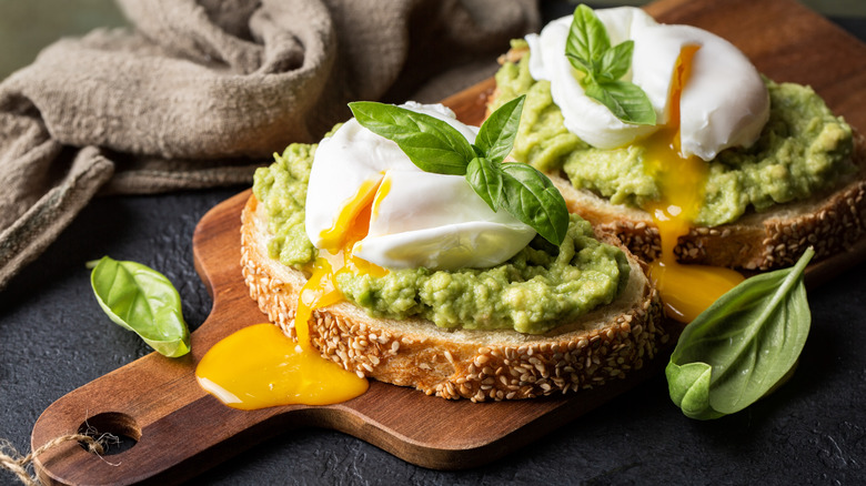 Two pieces of avocado toast on a cutting board, topped with with poached eggs and fresh basil.